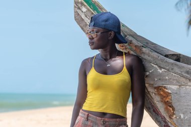 Güneş gözlüklü ve mavi şapkalı Afrikalı bir kadın Cape Coast Gana Batı Afrika 'daki eski bir balıkçı teknesine yaslanıyor..