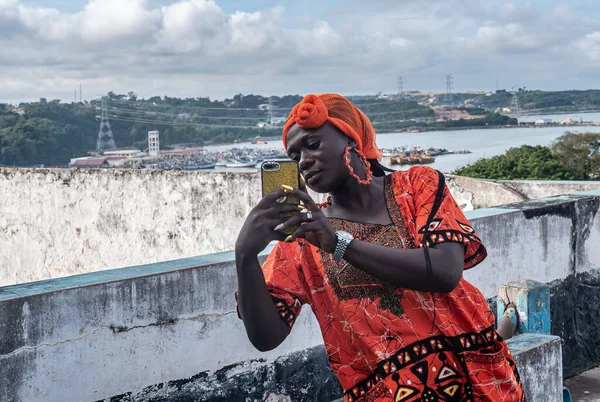 African woman with a mobile phone in hand and wearing traditional orange african dress. The place is Takoradi Ghana West Africa.