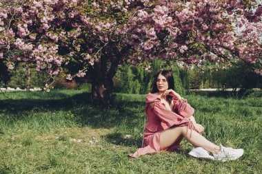 parkta genç bir bayan, kısa bir pembe yaz su birikintisi giymiş, büyük bir çiçek ağacı çiçeklenme ile çim üzerinde oturan