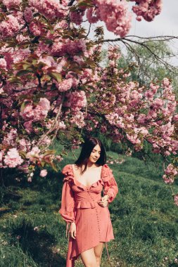 parkta genç bir esmer hizmetçi, güneş gözlüğü ile, bir çiçeklenme ağacı ile karıştırma, pembe ve seksi yazlık elbise giymiş