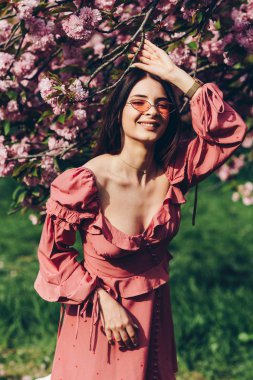 parkta genç bir esmer hizmetçi, güneş gözlüğü ile, bir çiçeklenme ağacı ile karıştırma, pembe ve seksi yazlık elbise giymiş
