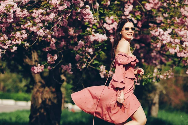 parkta genç bir esmer hizmetçi, güneş gözlüğü ile, bir çiçeklenme ağacı ile karıştırma, pembe ve seksi yazlık elbise giymiş