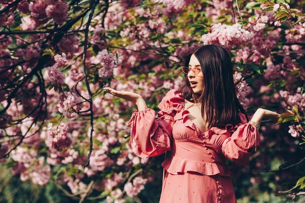 parkta genç bir esmer hizmetçi, kız düzeyinde açık avuç içi tutarak, ve şaşırır ve büyük bir pembe çiçeklenme ağacının arka planda şaşırır