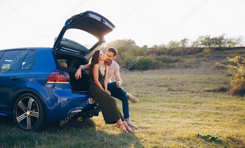 Happy young couple having a coffee break during road trip in countryside. Man and woman sitting in car trunk and having coffee.
