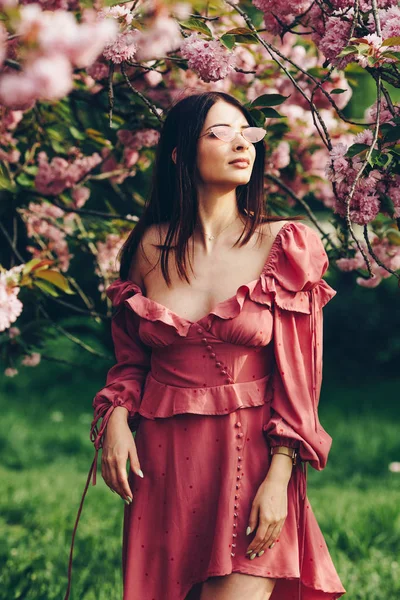 parkta genç bir esmer hizmetçi, güneş gözlüğü ile, bir çiçeklenme ağacı ile karıştırma, pembe ve seksi giymiş 