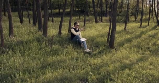 Düşünceli bir adam ormandaki çimlere oturur ve kitap okur. — Stok video