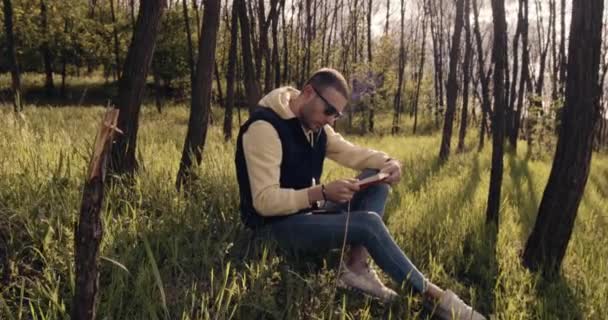 Düşünceli bir adam ormandaki çimlere oturur ve kitap okur. — Stok video