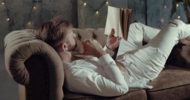 Sakallı bir adam kanepesinde uzanıyor ve kitap okuyor.