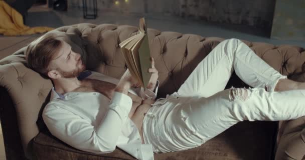 Bir adam kitap okurken kanepesinde uzanıyor. — Stok video