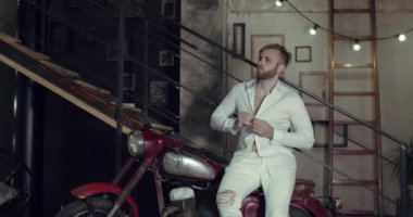 Yakışıklı bir adam oturuyor, sonra eski bir motosikletin üstünde dans ediyor.