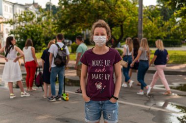 Maskeli genç bir kız sokakta kalabalığın içinde duruyor. Bir sürü insan maskeli geziyor.