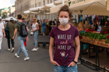 Maskeli genç bir kız sokakta kalabalığın içinde duruyor. Bir sürü insan maskeli geziyor.