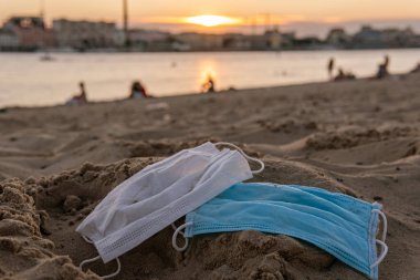 Kullanılan iki tek kullanımlık maske (COVID-19, Coronavirus) kumsalda kumla kaplıdır ve çevre kirliliği veya su kirliliği gibi kötü sonuçlara yol açar.