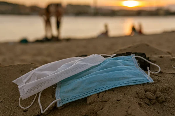 Two used disposable masks (protect from COVID-19, Coronavirus) are on a beach, covered with sand, leading to bad consequences like pollution or contamination of the nature and water