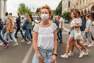Maskeli genç bir kız caddenin karşısındaki kalabalığın yanında duruyor. Bir sürü insan maskesiz yürüyor ve sosyal mesafeyi korumuyor.