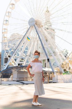 Sokakta maskeli genç bir kadın dönme dolabın yanındaki meydanda poz veriyor ve güneşli bir yaz gününde ciddi ciddi bakıyor.