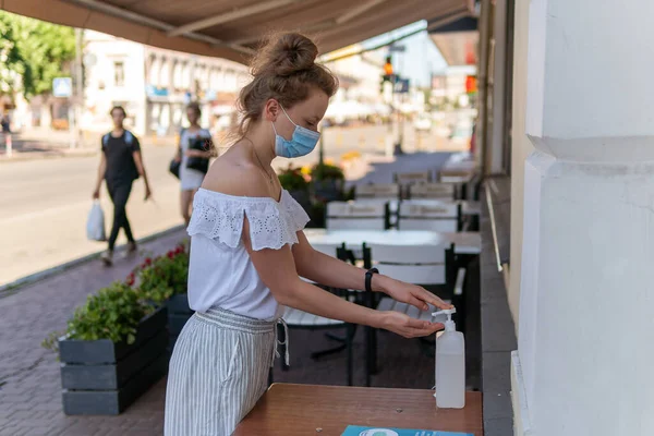Kyiv (Kiev), Ukrayna - 08 Ağustos 2020: Sokakta maske takmış güzel bir genç kadın, güneşli bir yaz gününde restoranın yakınında veya bir kafede antiseptik ile ellerini dezenfekte ediyor