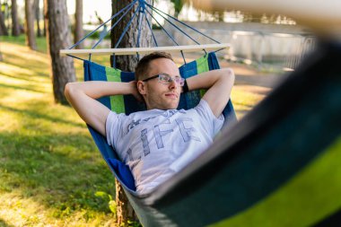 Gözlüklü çekici bir genç, güneşli bir günde, açık havada bir hamakta dinleniyor.