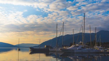 Ufukta büyük yatları ve tekneleri olan sessiz sakin bir liman. Dağlar ve bulutlar akşam alacakaranlıkta deniz suyuna yansıyor.
