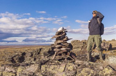 A man with a backpack on a hike stands on a mountain and looks into the distance. A man stands next to a pile of rocks cairn in the polar autumn tundra clipart