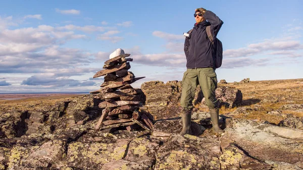 man with a backpack on a hike stands on a mountain and looks into the distance. A man stands next to a pile of rocks cairn