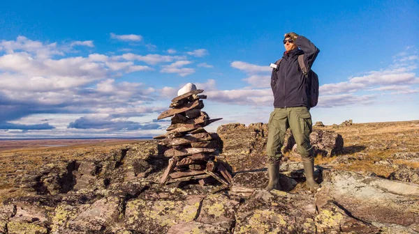 A man with a backpack on a hike stands on a mountain and looks into the distance. A man stands next to a pile of rocks cairn in the polar autumn tundra