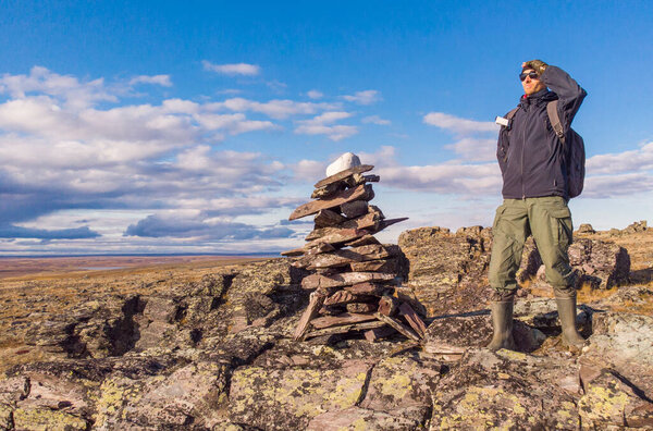 A man with a backpack on a hike stands on a mountain and looks into the distance. A man stands next to a pile of rocks cairn in the polar autumn tundra