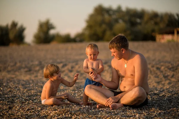 Familia feliz pasar tiempo juntos sentados en una playa de guijarros al atardecer . — Foto de Stock