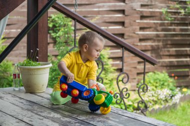 Sarı tişörtlü küçük bir çocuk evinin yakınında oyuncak bir traktör çalıyor.