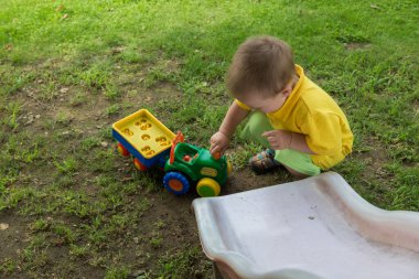 Sarı tişörtlü küçük bir çocuk evinin bahçesinde oyuncak bir traktörle oynuyor.