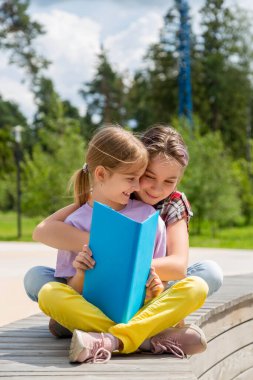 Dışarıda bankta oturmuş kitap okuyan iki genç kız. Bu kız kardeşler yaz boyunca ailecek vakit geçiriyorlar.