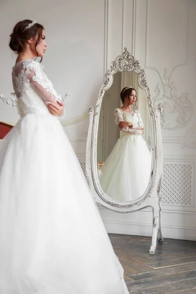 Full-length portrait of the bride in the reflection of the mirror. The bride is standing in a light studio, she has wedding makeup and styling, light from the window.