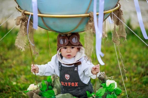 A little boy in a aviator\'s hat sits in a basket of a toy balloon in a field. High quality autumn art photography.