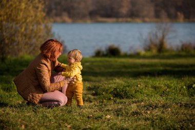 .Annesi sarı bir ceketle oğlunun yanına çömeldi. Sonbahar ormanı ve gölün arka planına yaslandı. Altın sonbahar Hint yazı.
