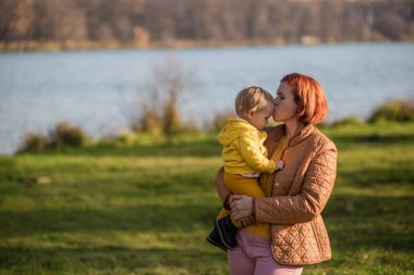 .Annem kucağında sarı bir ceketle küçük bir çocuğun oğlunu tutuyor ve onu alnından öpüyor. Sonbahar ormanı ve gölün arka planında, altın sonbahar Hindistan yazında.