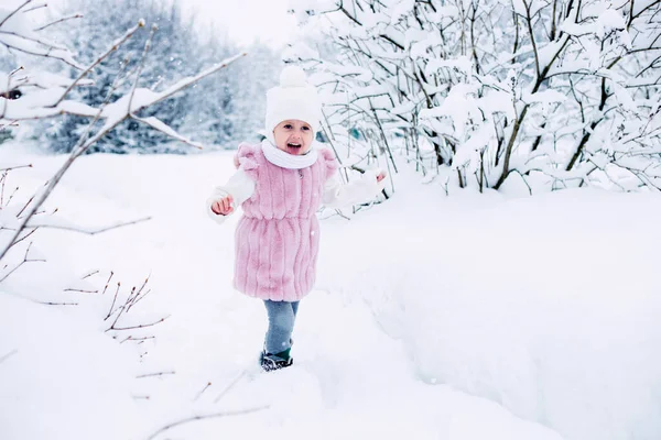 .A little girl in a pink fur coat stands in the middle of a snow-covered park on a cloudy winter day