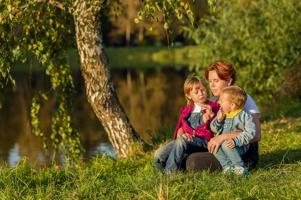 Familia feliz mamá y los niños se sientan en la orilla del río — Foto de Stock