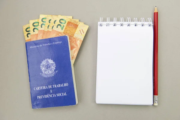 Çalışma kartının en üst görüntüsü, içinde para faturaları olan boş bir arkaplanda not defterinin yanında. Tercüme: Çalışma ve İstihdam Bakanlığı - İş ve Sosyal Güvenlik Kartı.