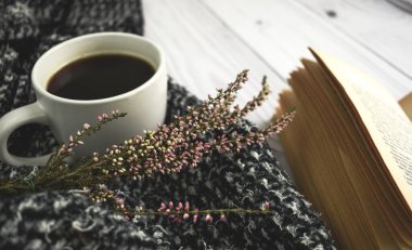 İçinde kitap, çiçek ve kırmızı sonbahar yaprağı olan bir fincan sade kahve ve kahverengi arka planda sıcak yün elbiseler. Düz konum, üst görünüm, kopyalama alanı. Sonbahar kompozisyonu, sonbahar konsepti.