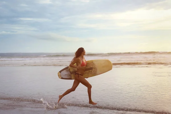 Een jonge vrouw met wit surfen in haar handen loopt langs de Oceaan kust bij zonsondergang. Stockfoto