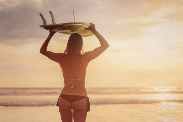 Een surfer met lang haar die terugstaat naar haar cel. Ze houdt een witte surfplank op haar hoofd bij zonsondergang. Surfer en Oceaan Rechtenvrije Stockfoto's