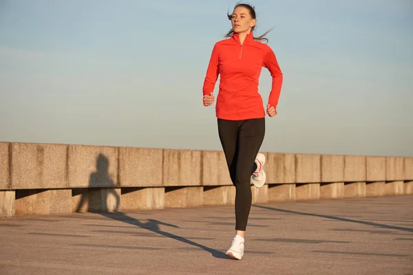 Een jogging vrouw in rood hardloopshirt en zwarte leggins op straat bij zonsopgang. Hardlopen op betonnen kade Stockafbeelding