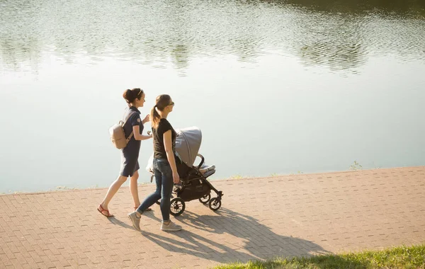 Moskova, Rusya - 26 Haziran 2020: Bebek arabasında bir bebekle gölün kenarındaki parkta yürüyen genç kadınlar. Yazın Moskova 'da gün batımında, anneler ve çocuklar, açık hava aktiviteleri.