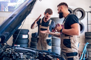 Üniformalı iki teknisyen otomobil servisinde çalışıyor. İletişim kontrolü ve onarım. Araba atölyesinde takım çalışması konsepti..