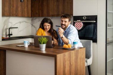 Bir çift evde rahat bir sabahı paylaşıyor, kahveyi yudumlarken ve ahşap aksanlı bir mutfakta hamur işlerinin tadını çıkarırken dizüstü bilgisayarla meşgul oluyorlar..