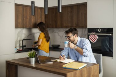 Mutfak adasında oturan bir adam kahve içerken deftere bir şeyler yazıyor. Bir kadın kahvaltı hazırlıyor, şık bir mutfakta rahat bir sabah ortamı yaratıyor..