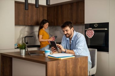 Bir adam şık bir mutfak adasında oturur, tabletine odaklanır, arka planda bir kadın ise yemek hazırlar. Mutfak pırıl pırıl ve çağdaş. Sıcaklık ve konfor saçıyor..