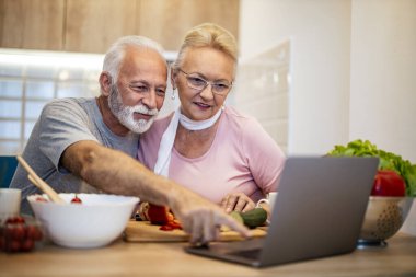 Yaşlı bir çift şık bir mutfakta lezzetli bir yemek pişirme deneyimi yaşıyor, yemek tarifi fikirleri için dizüstü bilgisayara bakarken aynı zamanda taze malzemeler hazırlıyor..