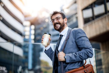 Kendine güvenen bir işadamı, çağdaş ofis binalarının yanında yürürken kahve fincanı tutuyor ve gülümsüyor. Parlak güneş ışığı şehir ortamının canlı atmosferini arttırır..