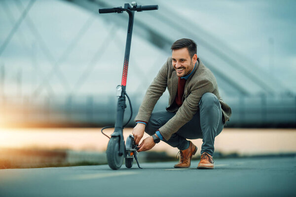 Улыбающийся мужчина в пиджаке и туфлях на коленях, чтобы настроить свой электрический скутер на городской тропе. Мягкий свет заката подчеркивает окружающие городские пейзажи.